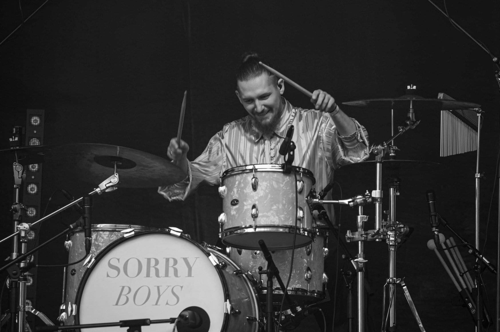 Uśmiechnięty perkusista w koszuli w paski gra na perkusji z napisem 'Sorry Boys' w czarno-białym ujęciu koncertowym.
