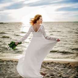 Mfilm Media - Panna młoda w zwiewnej sukni, z bukietem białych róż, stoi na piaszczystej plaży, spoglądając w stronę morza o zachodzie słońca.