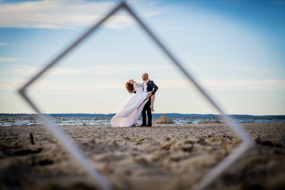 Para w tańcu na plaży, uchwycona przez ramę. Panna młoda w zwiewnej sukni, pan młody w garniturze. W tle morze i błękitne niebo. Nietypowe ujęcie, piasek na pierwszym planie.