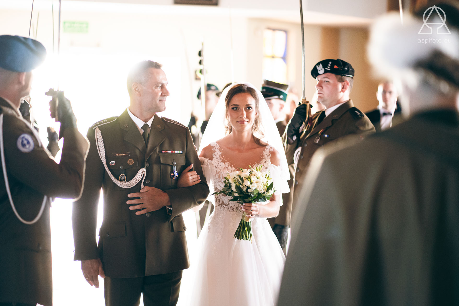Panna młoda w białej sukni prowadzona do ołtarza przez mężczyznę w mundurze wojskowym, otoczona przez żołnierzy z szablami. Uroczystość ślubna w kościele.