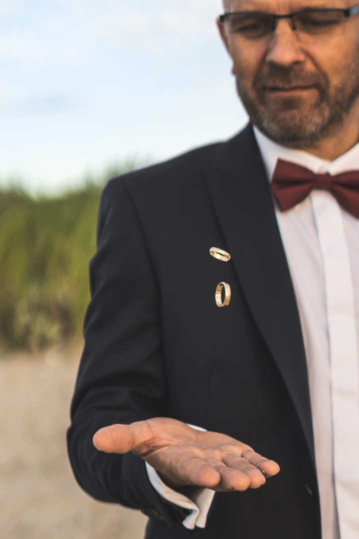 Elegancki mężczyzna w garniturze z muszką prezentuje dwa unoszące się w powietrzu złote obrączki ślubne na tle plaży i zieleni.