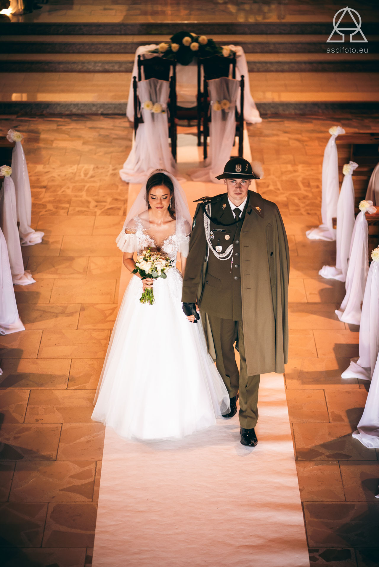 Para młoda w kościele: panna młoda w białej sukni z bukietem, pan młody w mundurze. Ślubna fotografia z uroczystości w stonowanych barwach, wykonana z góry.