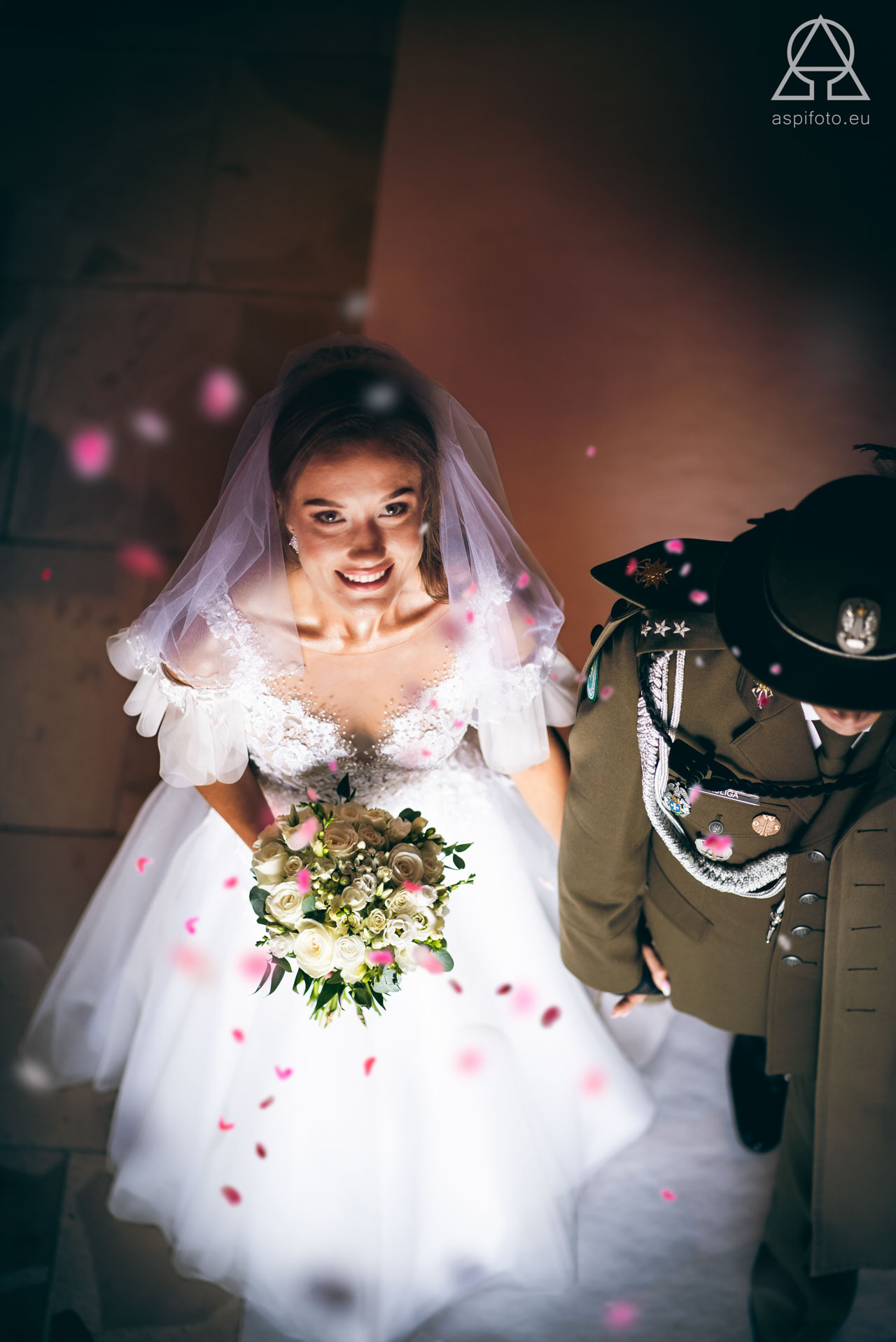 Panna młoda w białej sukni spogląda w górę, trzymając bukiet. Obok stoi mężczyzna w mundurze wojskowym. Widok z góry, płatki róż opadają.