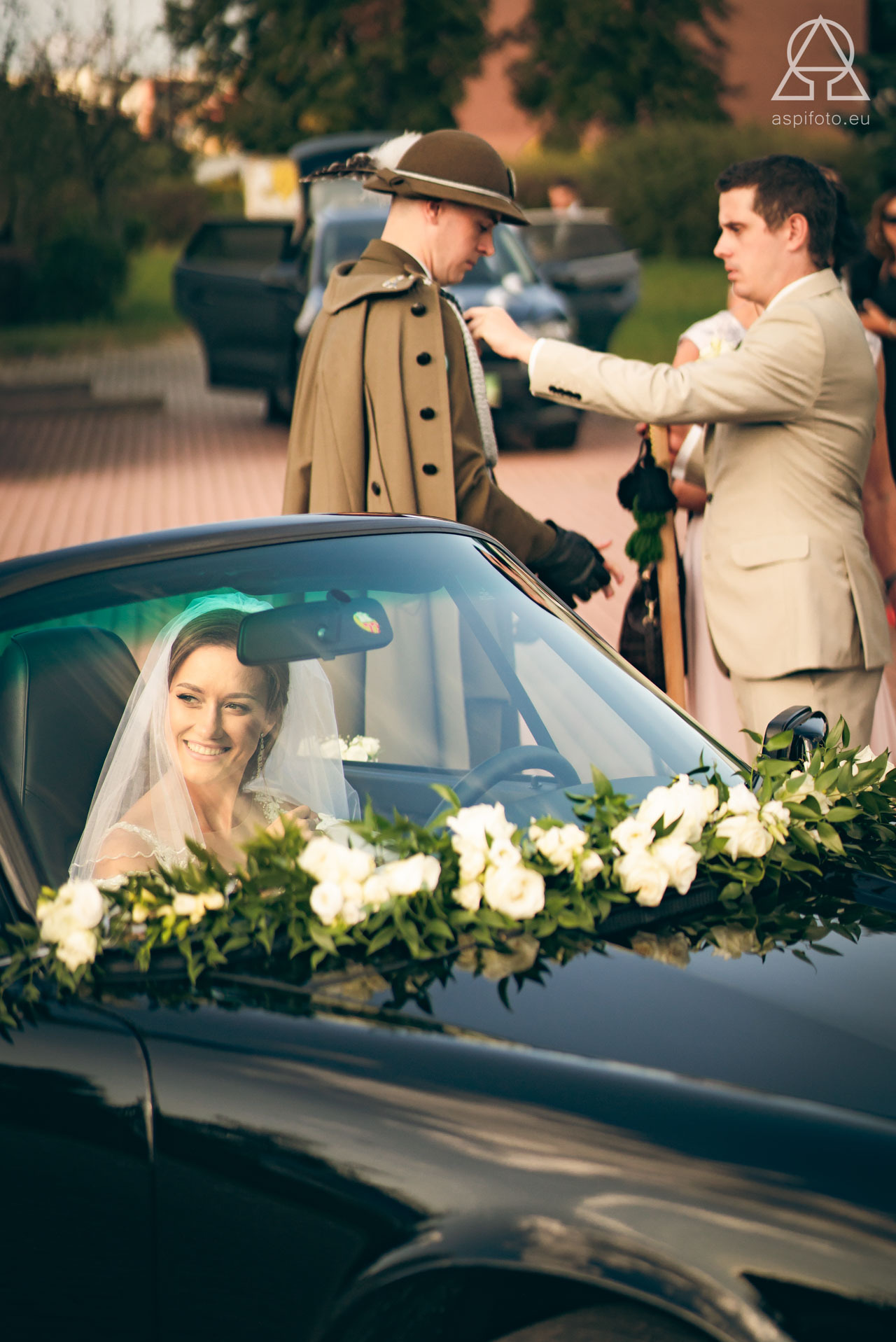 Uśmiechnięta panna młoda w samochodzie ozdobionym białymi kwiatami, w tle pan młody i drużba w tradycyjnym stroju. Nietypowe ujęcie z wesela.