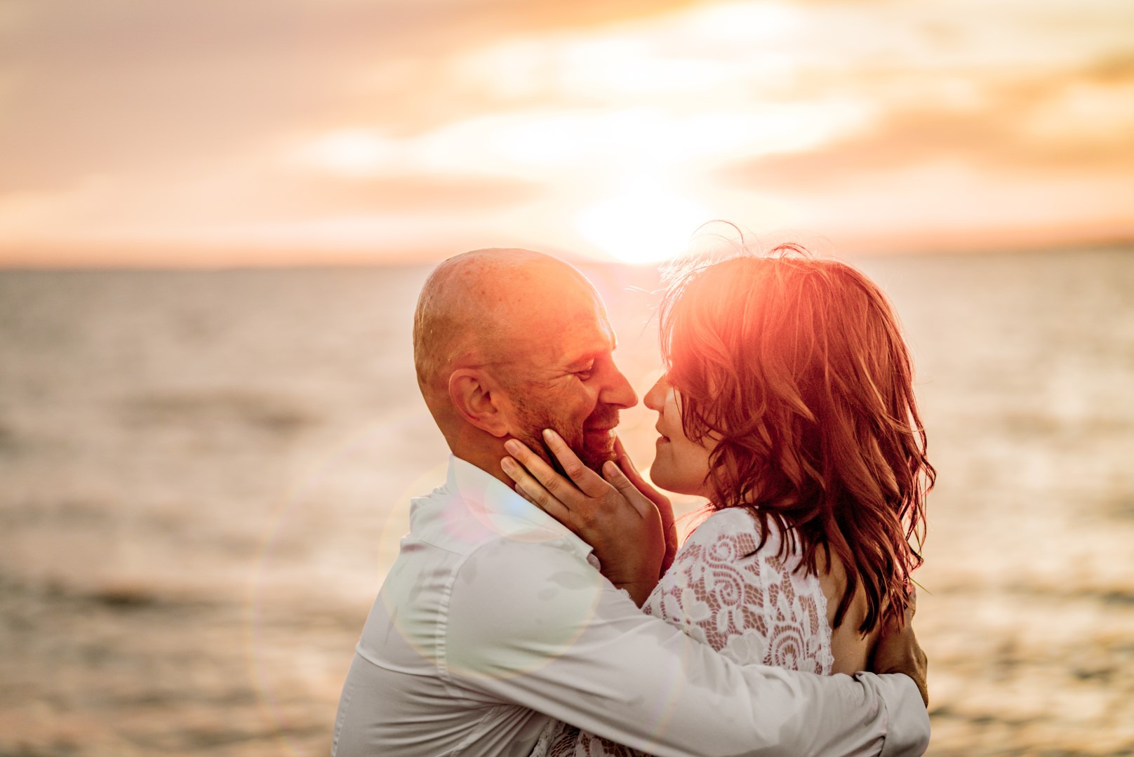 Para zakochanych na tle zachodzącego słońca nad morzem. Kobieta obejmuje mężczyznę, dotykając jego twarzy. Romantyczna sceneria, ciepłe światło.