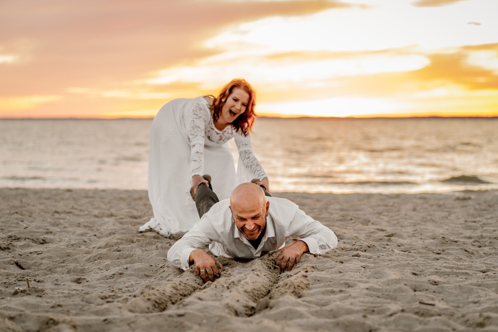 Radosna panna młoda ciągnie pana młodego po piasku na tle zachodzącego słońca. Nietypowa sesja ślubna na plaży.
