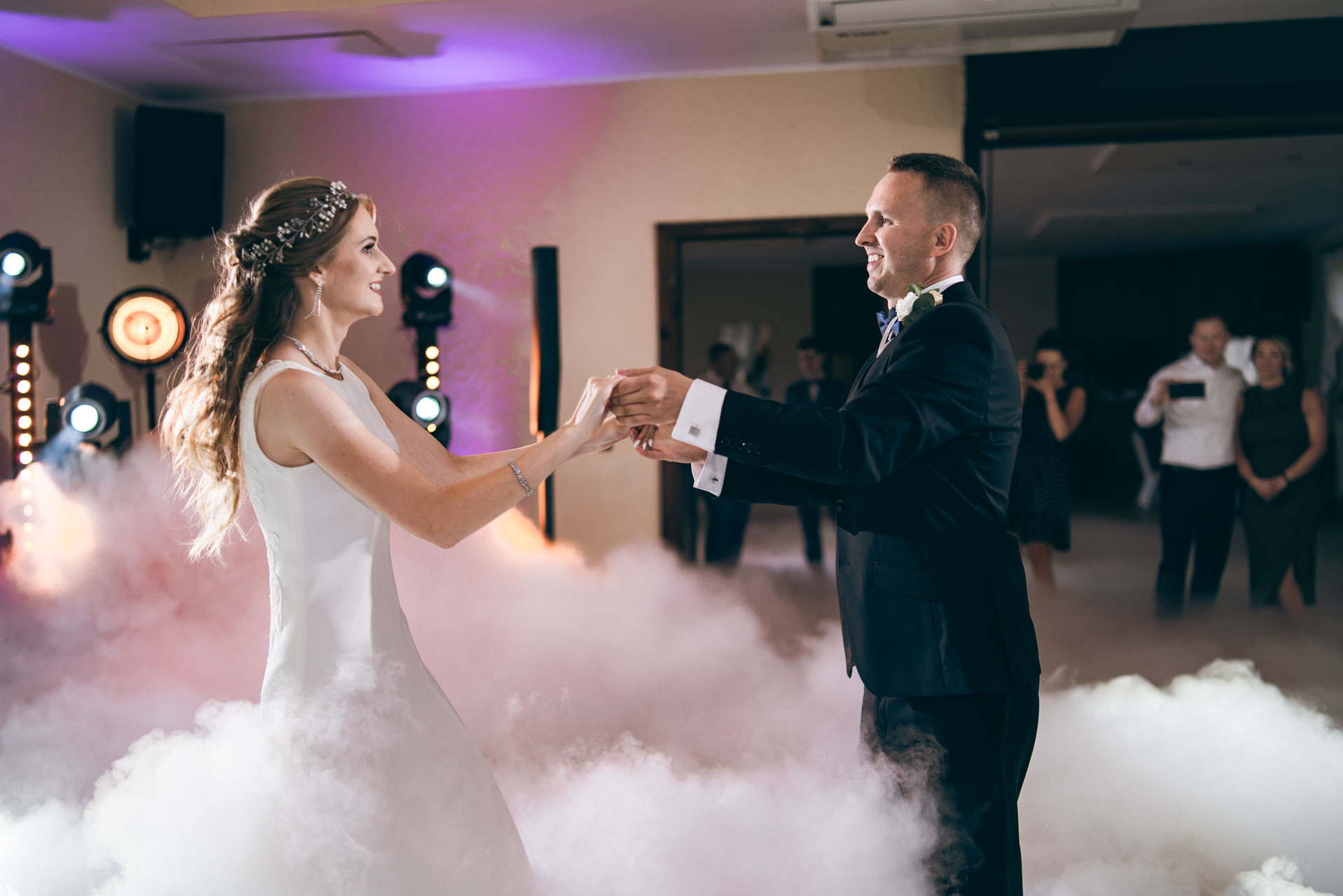 Uśmiechnięta para młoda w tańcu, otoczona efektem mgły, na tle oświetlenia scenicznego i obserwujących gości, podczas uroczystości ślubnej.