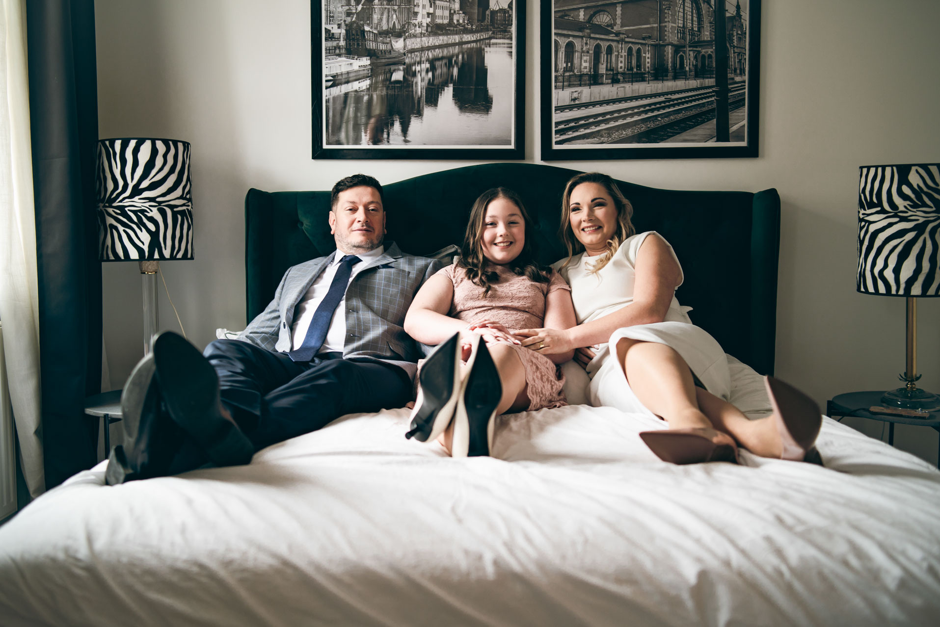 Uśmiechnięta rodzina (ojciec, córka i matka) w eleganckich strojach pozuje na łóżku z zielonym zagłówkiem, w tle lampy w zebrę i zdjęcia budynków.
