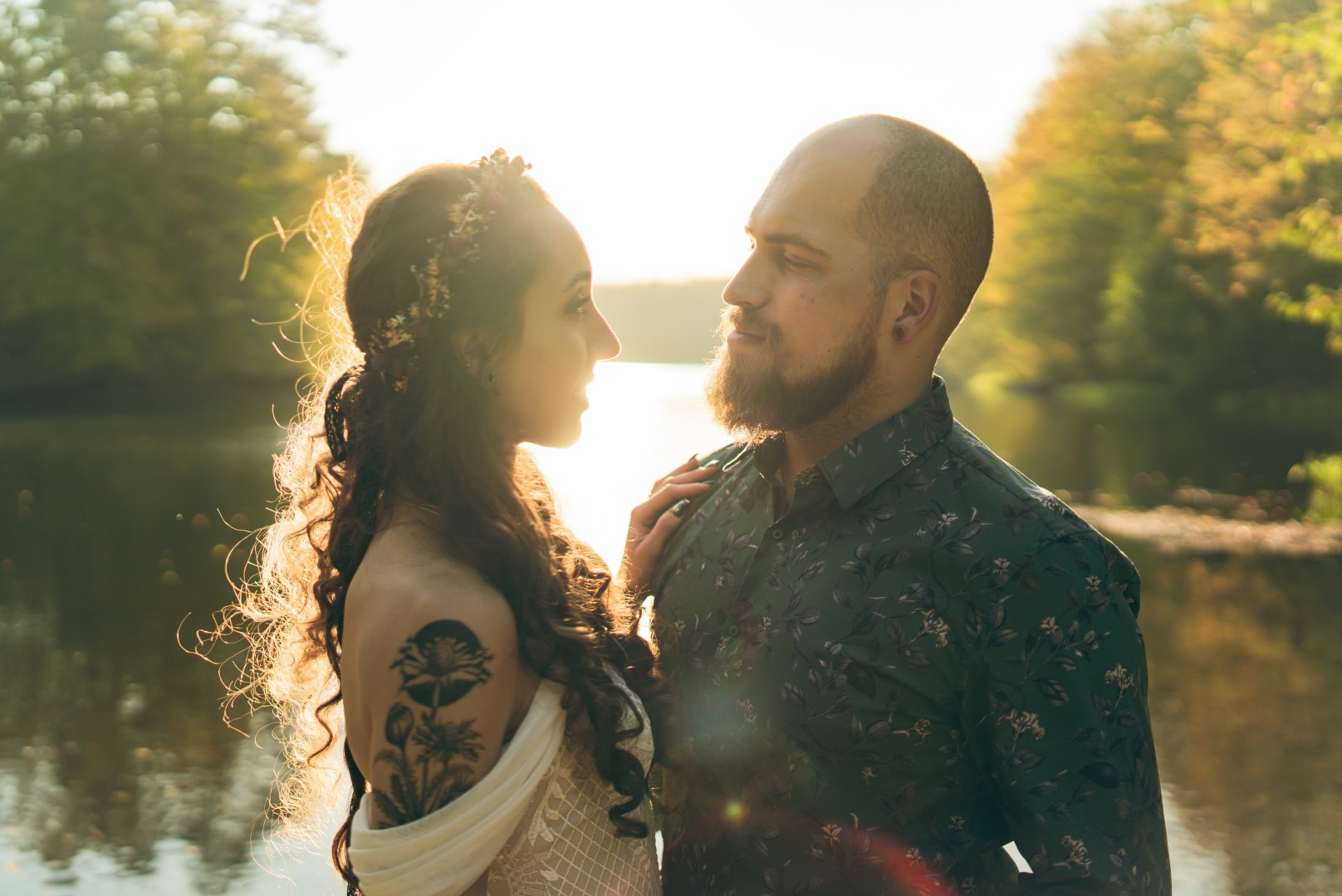 Zakochana para w złotym świetle, ona z kwiatami we włosach i tatuażem, on w koszuli w kwiaty, jezioro w tle, uchwycone w ciepłej, intymnej atmosferze.