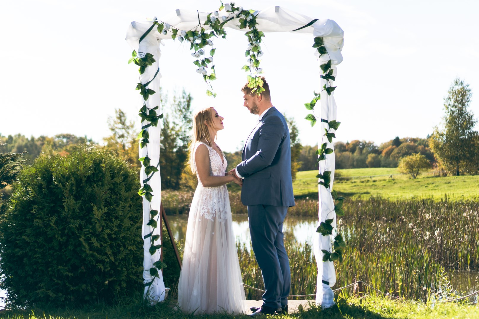 Para młoda trzymająca się za ręce pod zdobionym kwiatami i zielenią baldachimem na tle malowniczego krajobrazu z jeziorem i drzewami w słoneczny dzień.