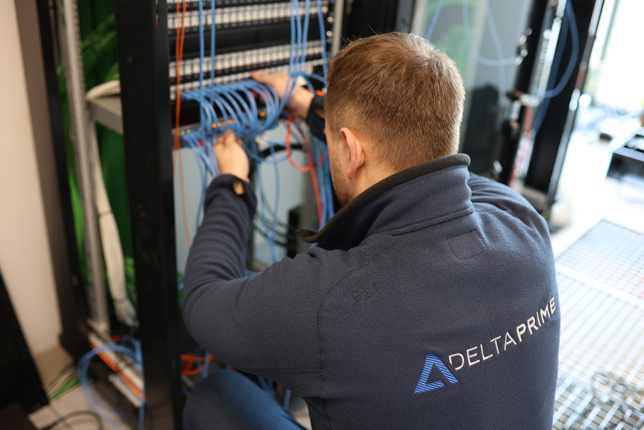 Instalator w bluzie Delta Prime łączy kable w szafie serwerowej. Widoczne niebieskie przewody i elementy szafy RACK.