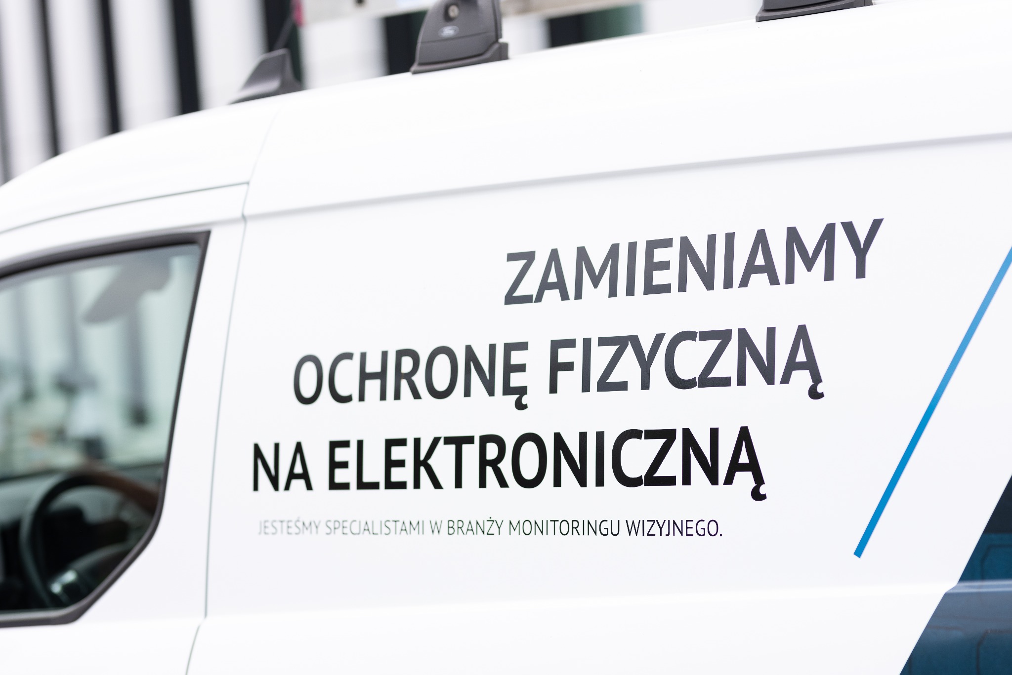 Biały samochód dostawczy z napisem: Zamieniamy ochronę fizyczną na elektroniczną. Jesteśmy specjalistami w branży monitoringu wizyjnego.