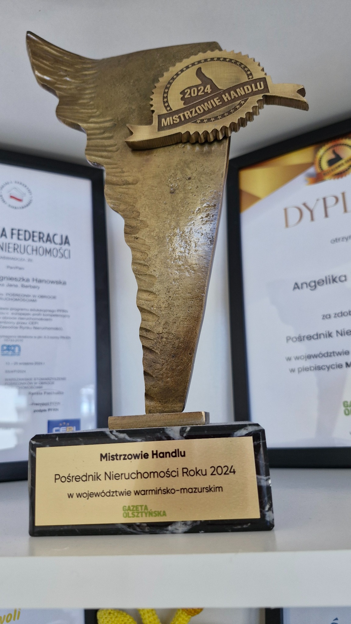 Statuetka 'Mistrzowie Handlu - Pośrednik Nieruchomości Roku 2024', z dyplomami w tle, symbolizująca sukces w branży. Nagroda z napisem 'Gazeta Olsztyńska'.