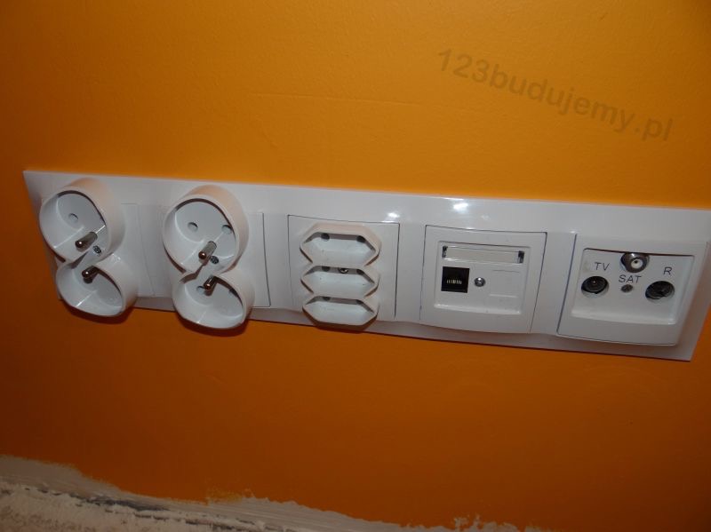 Biały panel z gniazdkami: podwójne gniazdo elektryczne, potrójne gniazdo, gniazdo internetowe, gniazdo TV/SAT/R, na pomarańczowej ścianie. Widok z dołu.