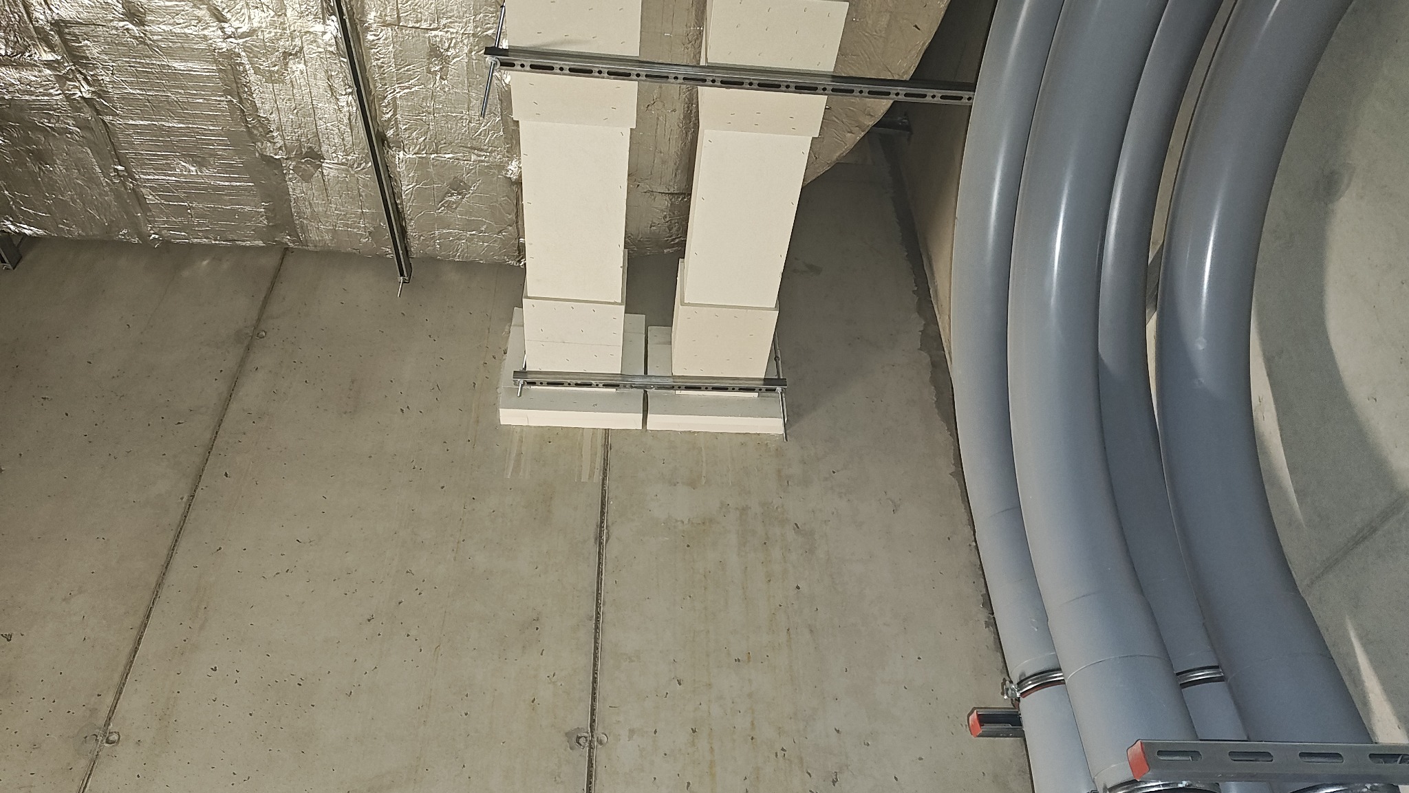 Instalacja wentylacyjna z izolacją i rury w szarym kolorze zamocowane do sufitu w surowym stanie. Widoczne elementy montażowe.