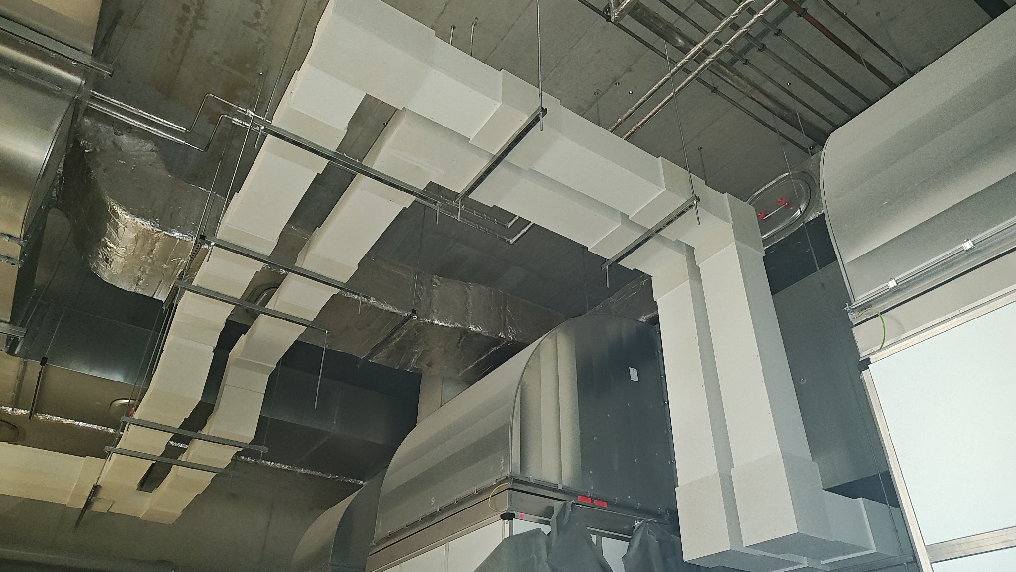 Widok na sufit z instalacją wentylacyjną, rury i kable prowadzone pod stropem. Elementy w kolorze srebrnym i białym, oświetlenie sztuczne.