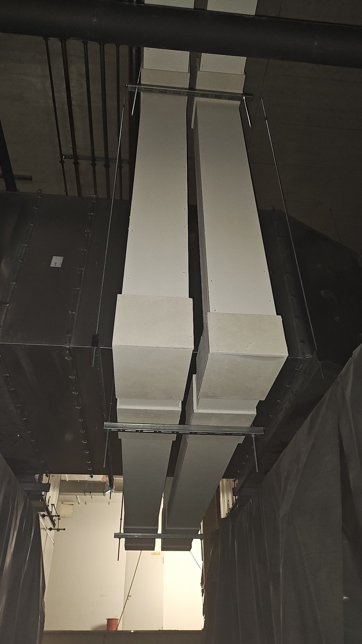 Dwa pionowe, prostokątne słupy z płyt karton-gips, przymocowane do sufitu za pomocą metalowych wsporników. Widoczne rury i instalacje pod sufitem. Ujęcie z dołu.