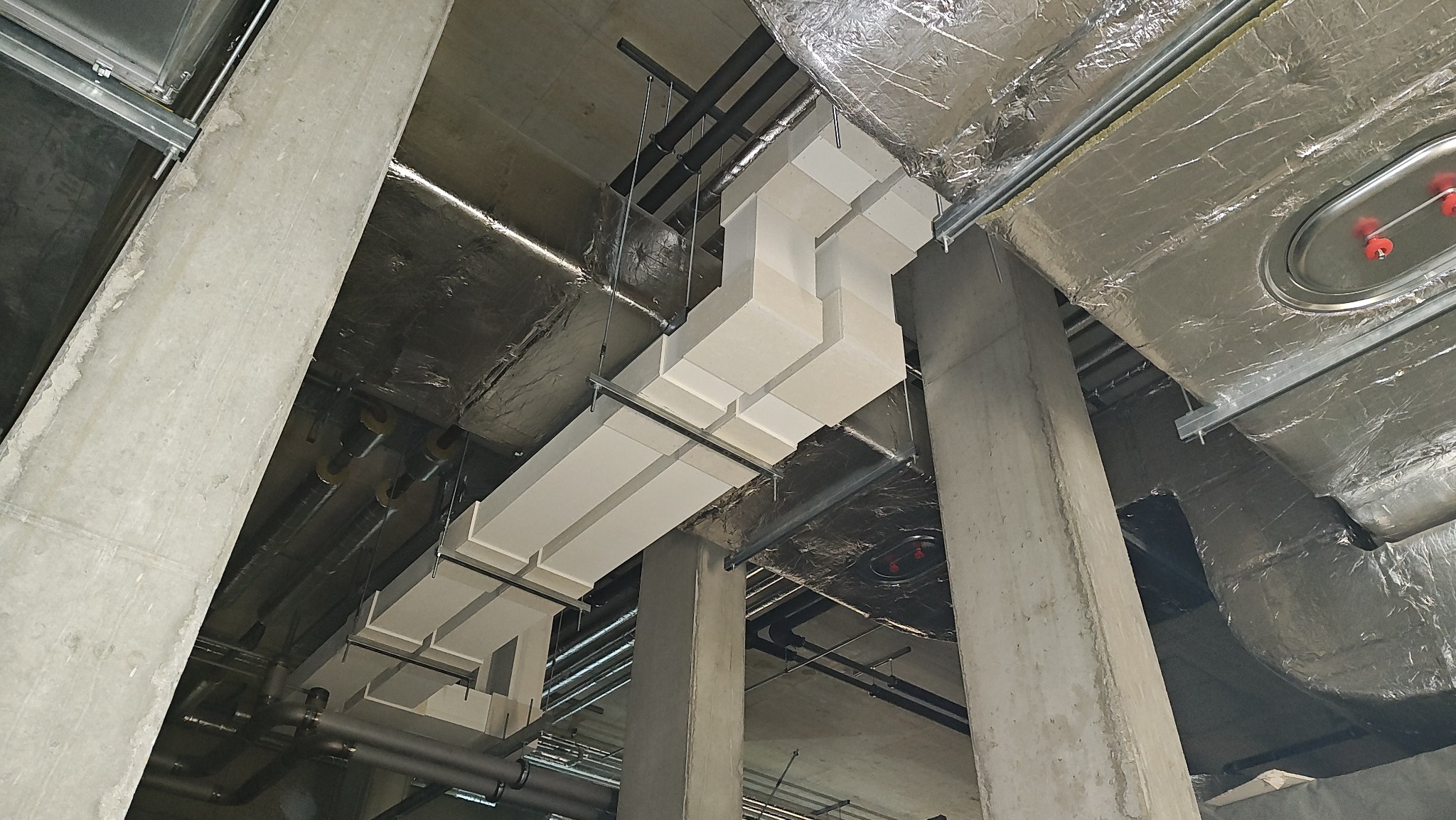 Instalacja wentylacyjna z białych, prostokątnych kanałów, zawieszona pod sufitem w surowym, przemysłowym wnętrzu. Widoczne betonowe kolumny i izolacja termiczna.