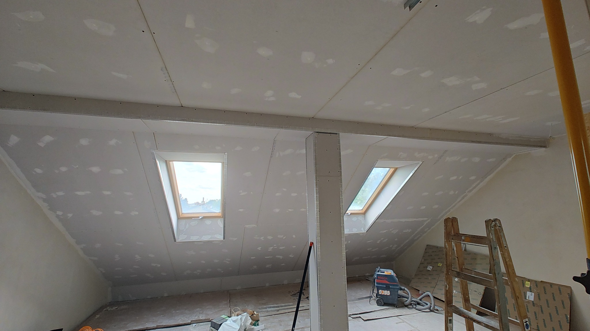 Poddasze z oknami dachowymi w trakcie montażu płyt kartonowo-gipsowych, widoczne szpachlowane łączenia, drabina i odkurzacz przemysłowy w pomieszczeniu.