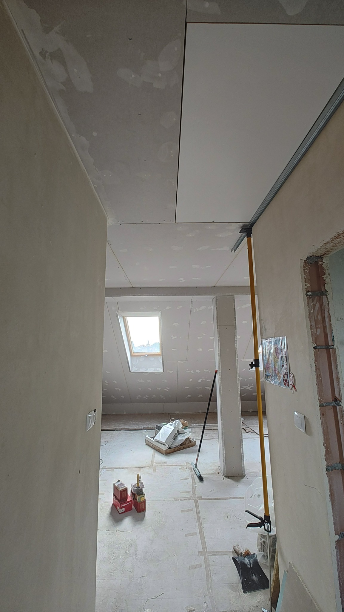 Montaż płyt karton-gips na suficie poddasza, widoczne profile i narzędzia. Częściowo wykończone ściany i sufit, okno dachowe w tle.