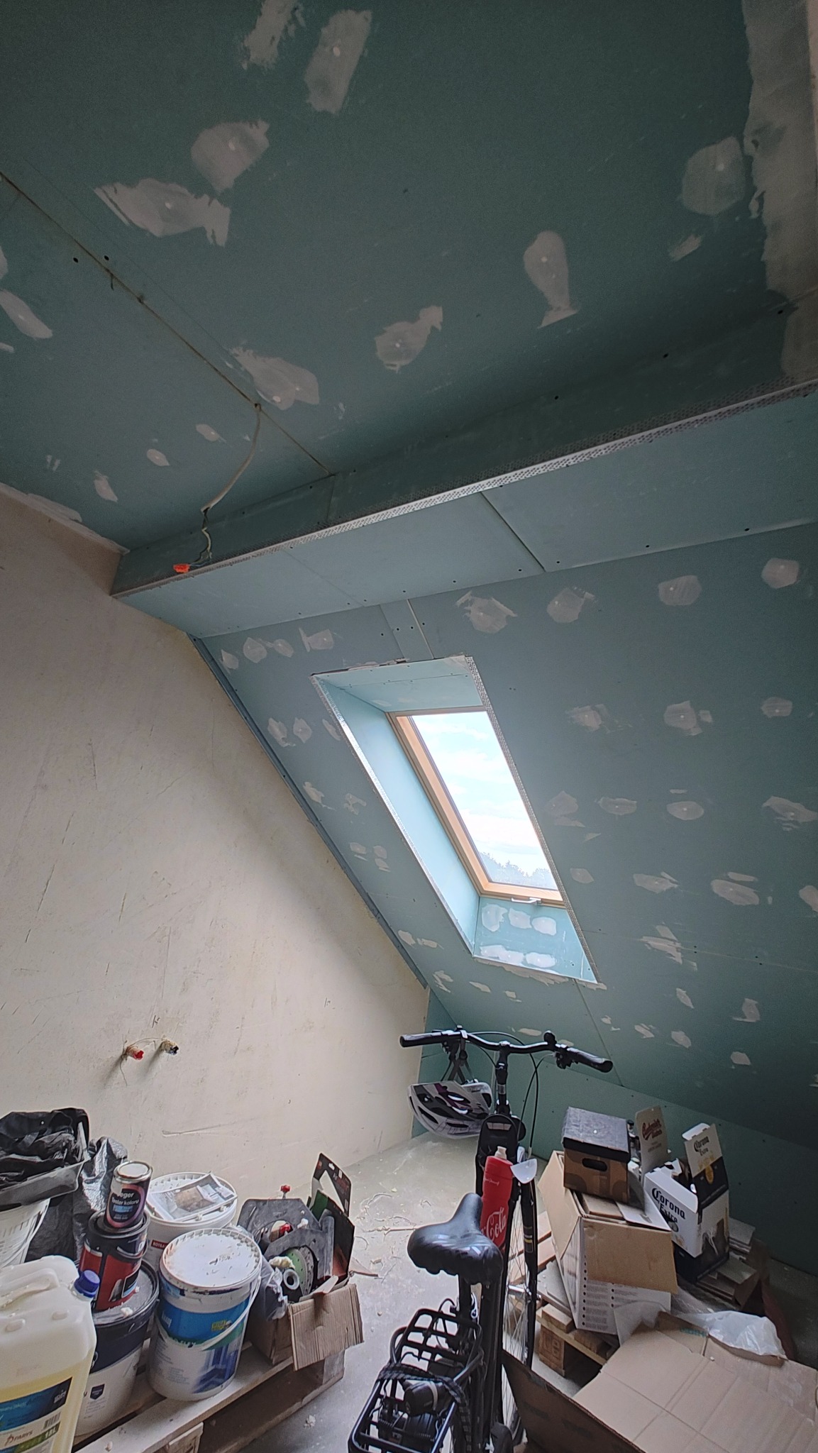 Poddasze w trakcie prac wykończeniowych: zamontowana płyta karton-gips na skosach, widoczne szpachlowane łączenia, okno dachowe. Na podłodze farby i rower.