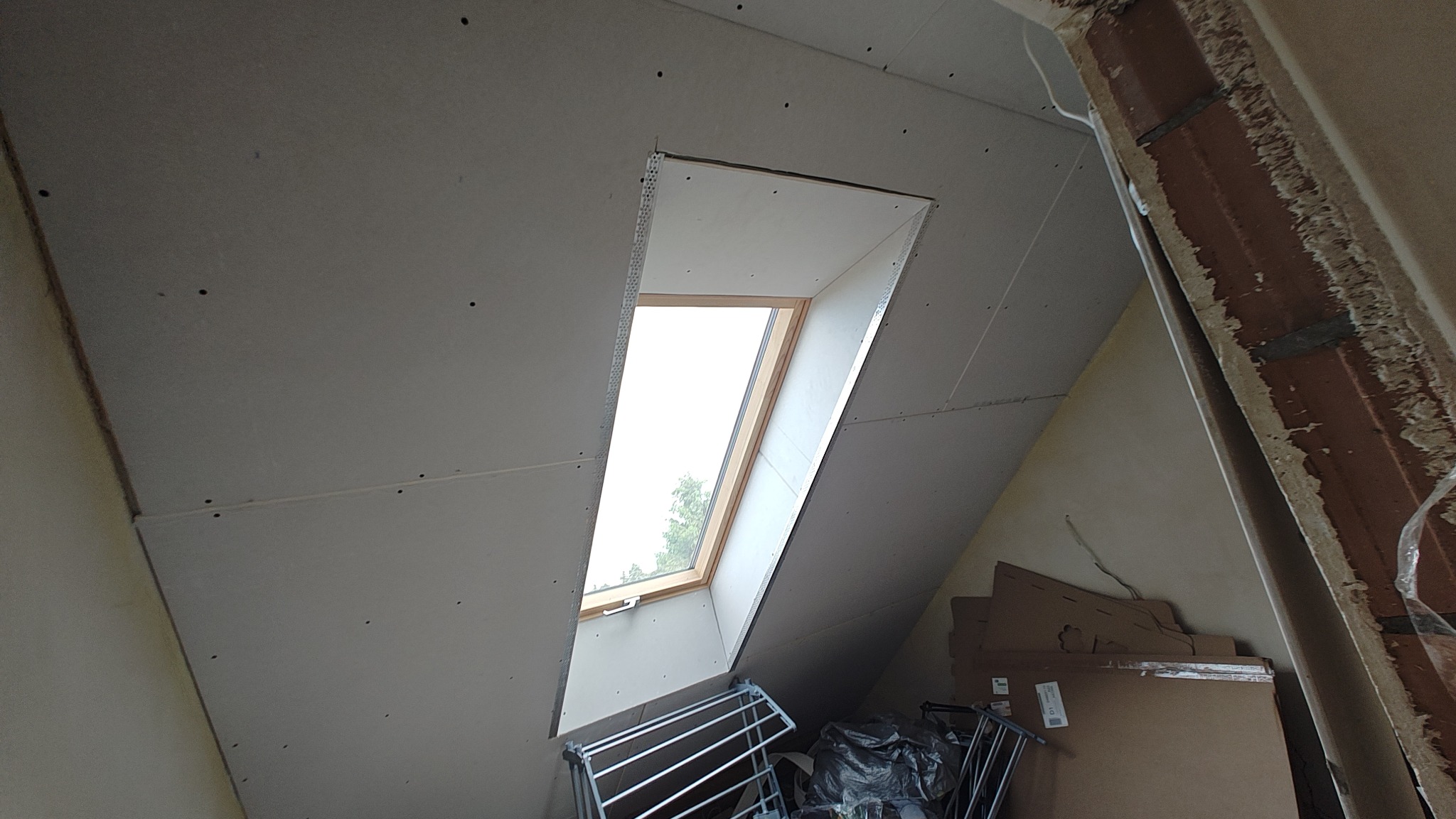 Montaż płyty KG wokół okna dachowego na poddaszu. Widoczne łączenia płyt, metalowe profile okienne i fragmenty konstrukcji budynku. Prace wykończeniowe w toku.