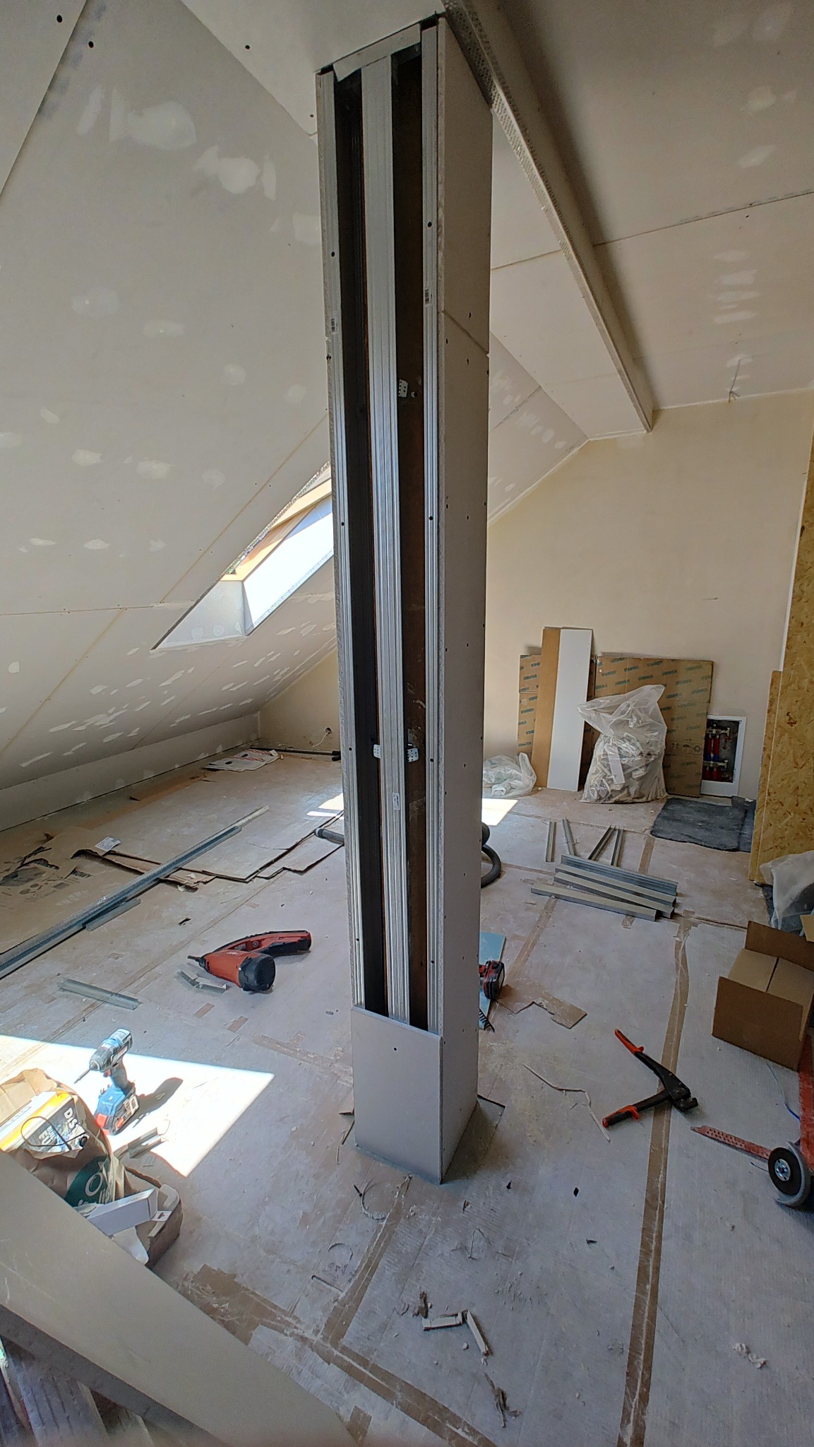 Montaż słupa z profili w poddaszu z oknem połaciowym. Widoczne narzędzia i płyty G-K na ścianach. Etap prac wykończeniowych w jasnym pomieszczeniu.