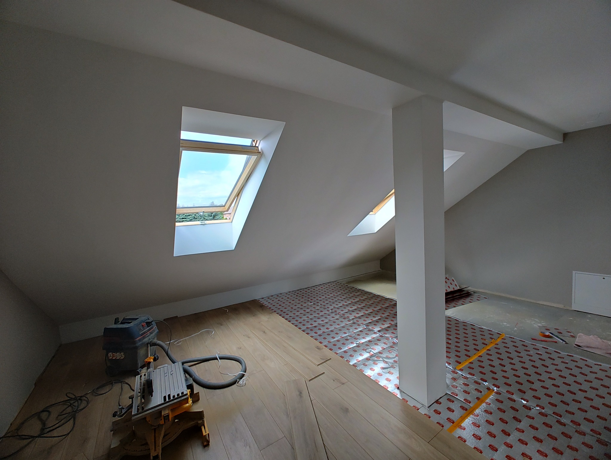 Poddasze z oknami dachowymi w trakcie układania paneli podłogowych. Widoczny parkiet, podkład izolacyjny, narzędzia i odkurzacz przemysłowy.