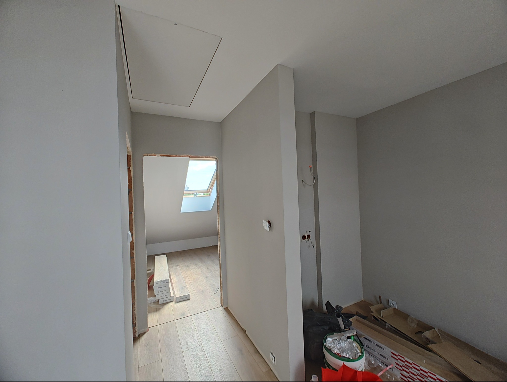 Wnętrze w trakcie remontu: szare ściany, widoczne kable, podłoga z paneli, okno dachowe. Na podłodze leżą panele i narzędzia. Widoczny otwór w suficie.