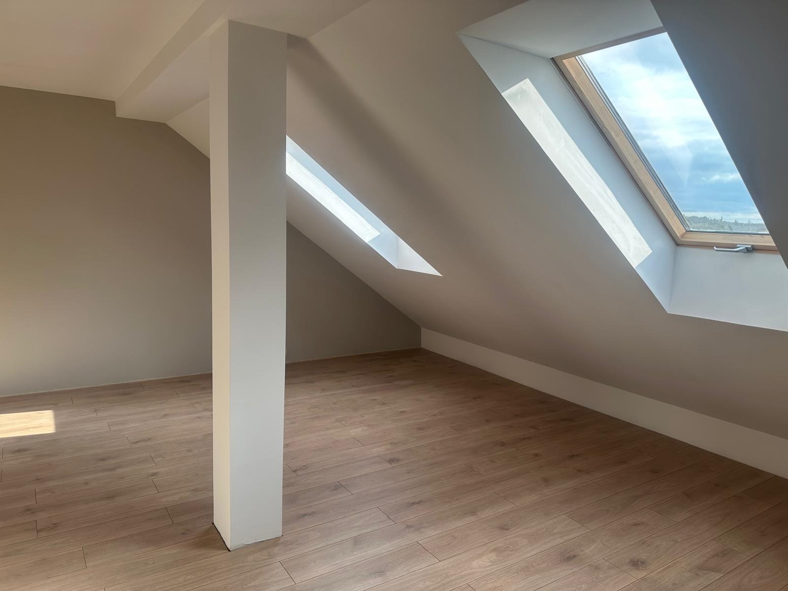 Jasne poddasze po remoncie: drewniane panele podłogowe, białe ściany i filar, okna dachowe wpuszczają naturalne światło. Nowoczesne wykończenie wnętrz.