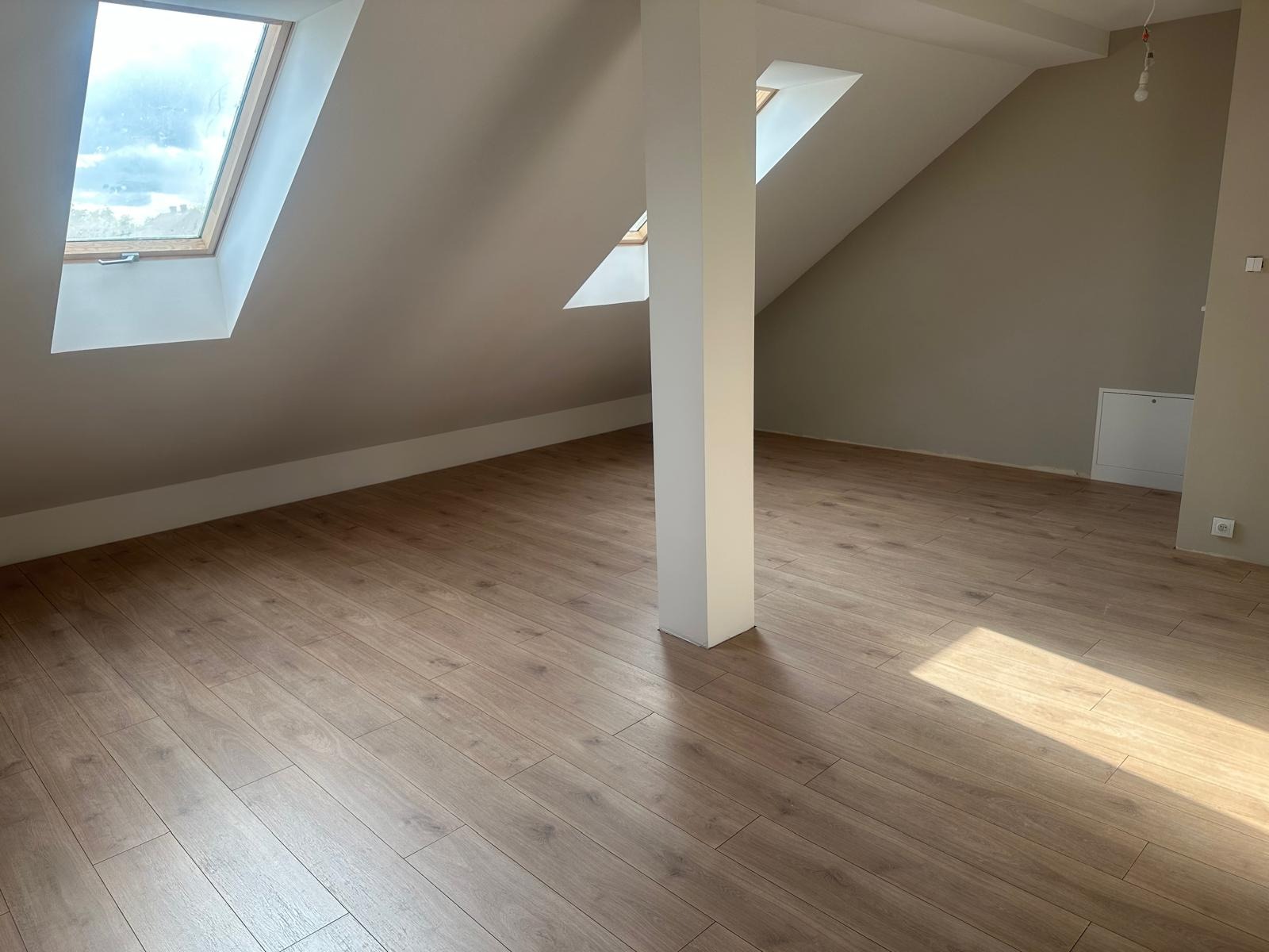 Podłoga z paneli w jasnym odcieniu drewna w pokoju na poddaszu z oknami dachowymi i kolumną nośną. Widoczne łączenia paneli i delikatne światło słoneczne.