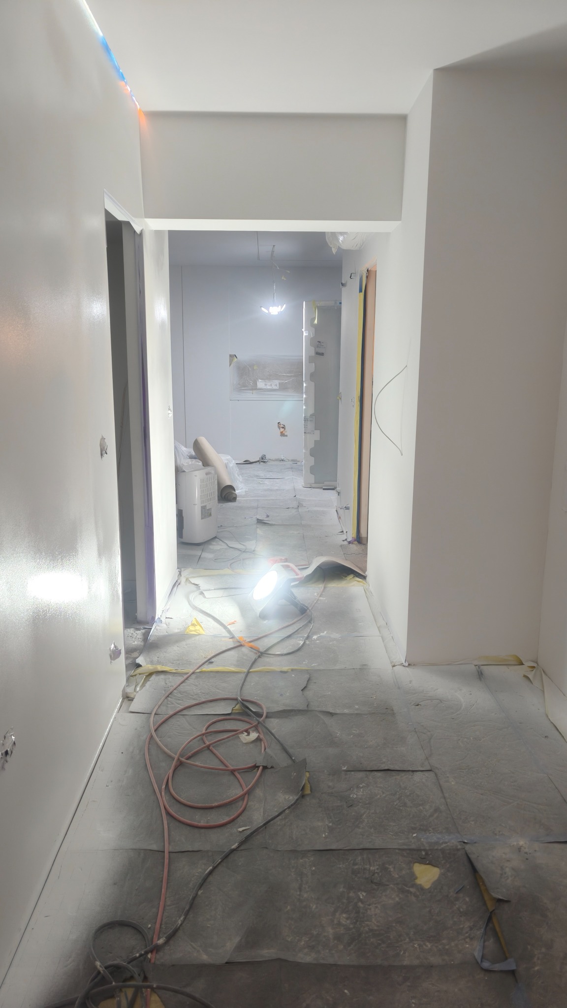 Korytarz w trakcie malowania, podłoga zabezpieczona folią, widoczne narzędzia i lampa halogenowa. Świeżo pomalowane ściany, w tle rolka folii malarskiej i osuszacz.