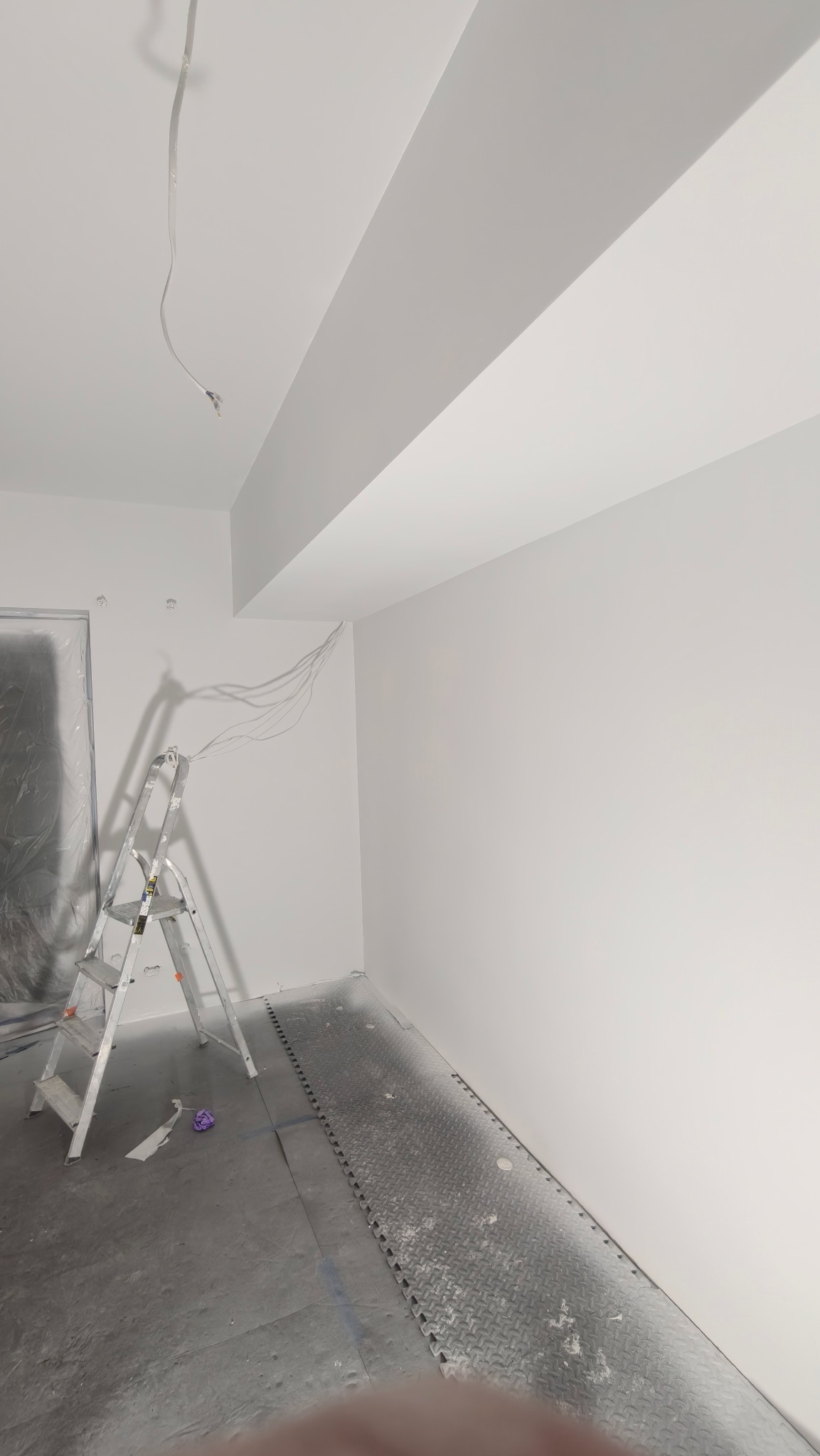 Białe ściany po malowaniu w pokoju z drabiną i zabezpieczoną folią podłogą. Widoczne kable wystające ze ściany.
