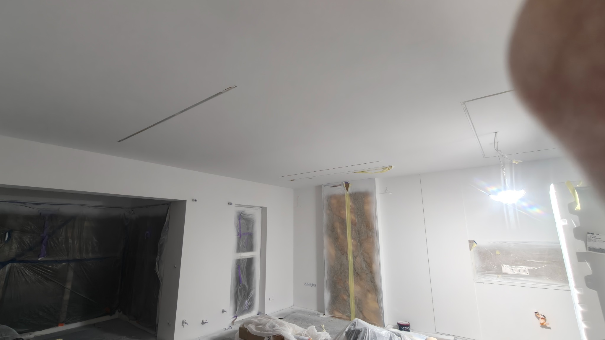 Białe wnętrze w trakcie prac wykończeniowych: okna i drzwi zabezpieczone folią malarską, widoczne listwy oświetleniowe na suficie. Przygotowanie do malowania.