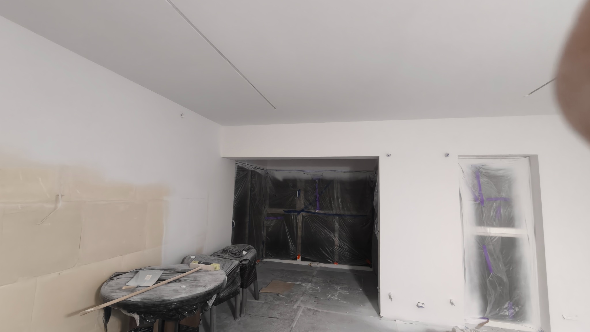 Wnętrze w trakcie prac wykończeniowych: ściany po szpachlowaniu, zabezpieczone folią okna i przejścia, narzędzia na stole. Widoczne przygotowania do malowania.