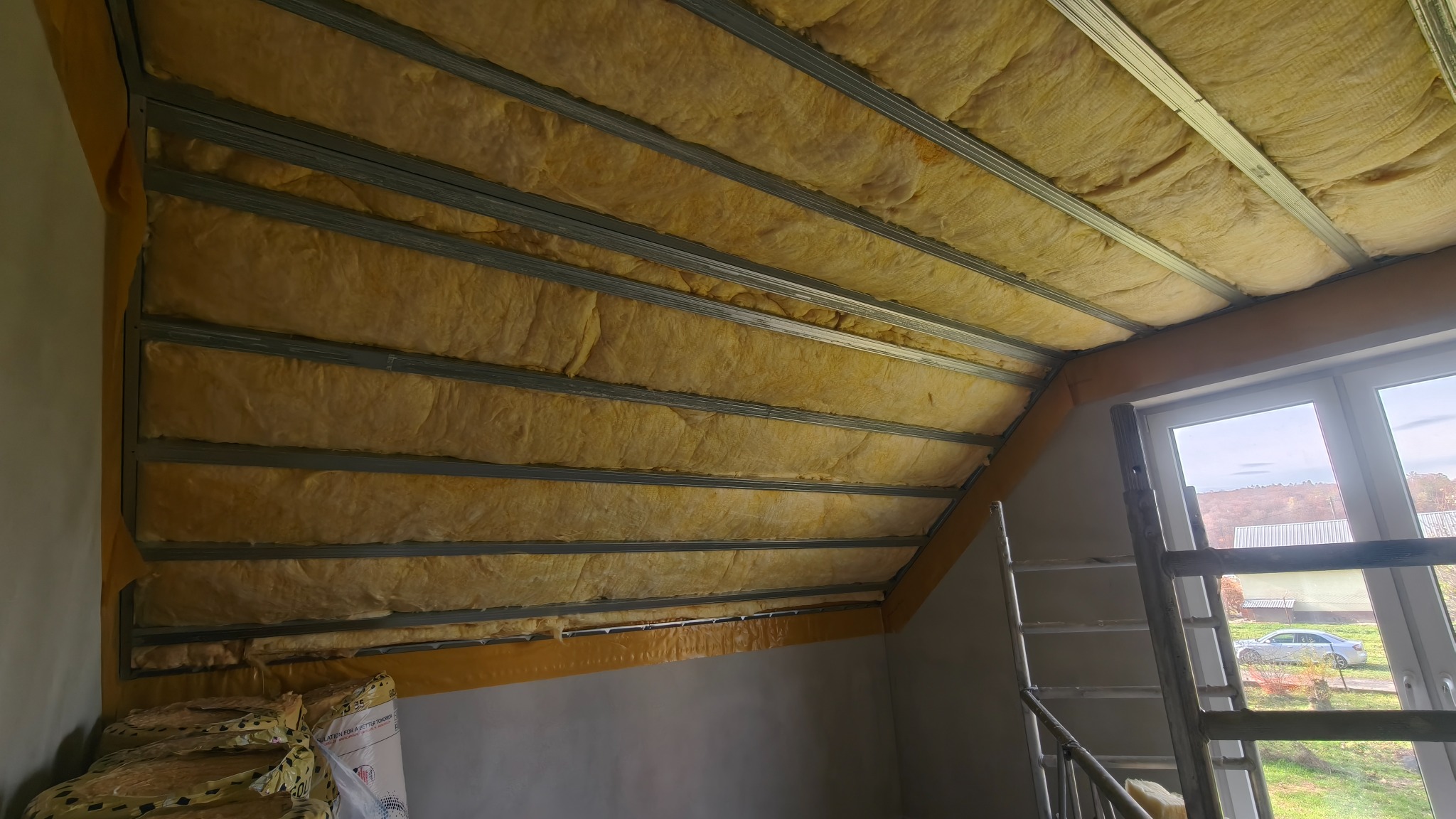 Skośny sufit poddasza z izolacją z wełny mineralnej, widoczne metalowe profile konstrukcyjne, okno w tle. Prace wykończeniowe na etapie ocieplania.