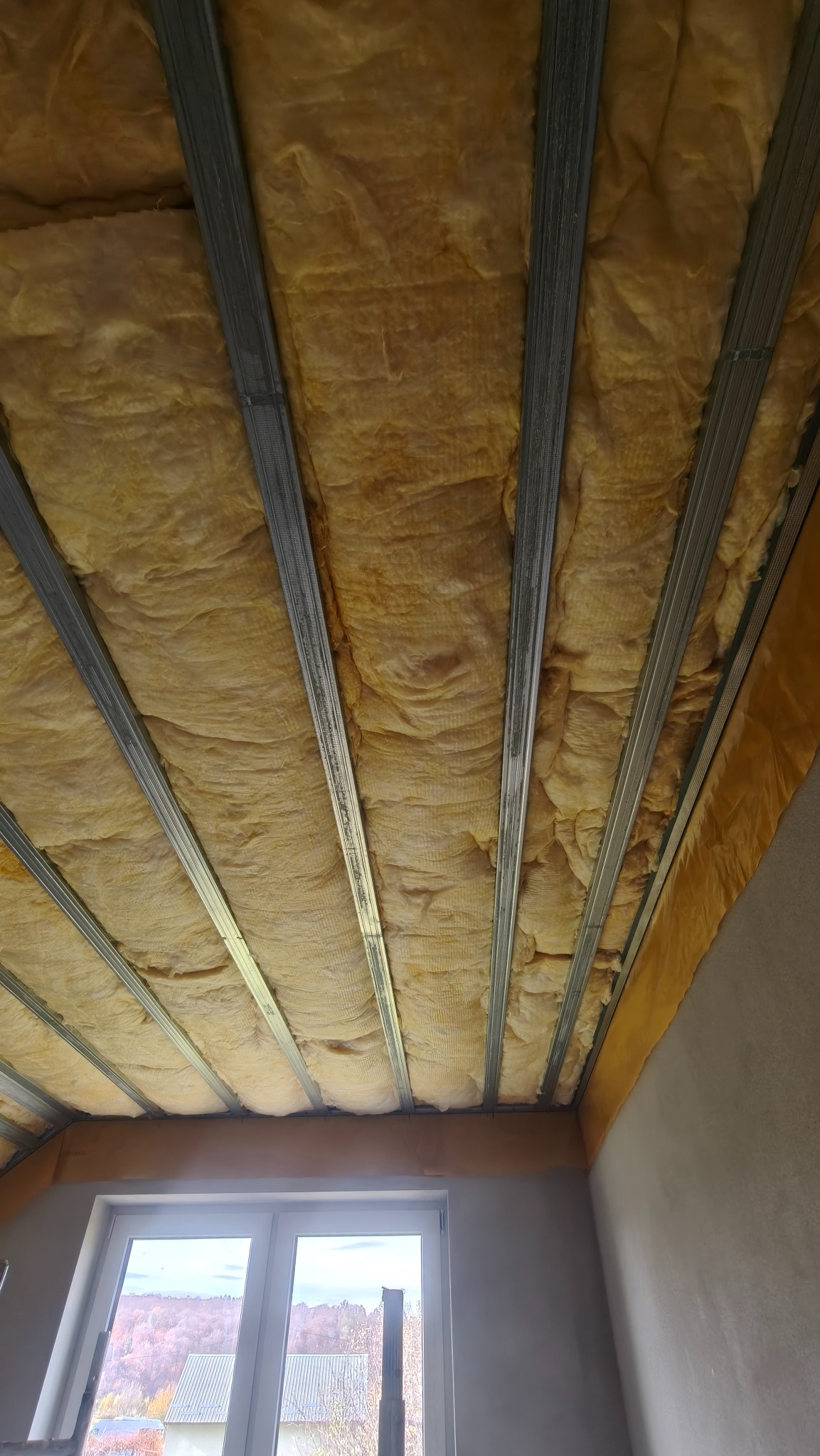 Sufit podwieszany z izolacją z wełny mineralnej, widok z dołu na metalową konstrukcję i ocieplenie, okno w tle. Prace wykończeniowe na poddaszu.