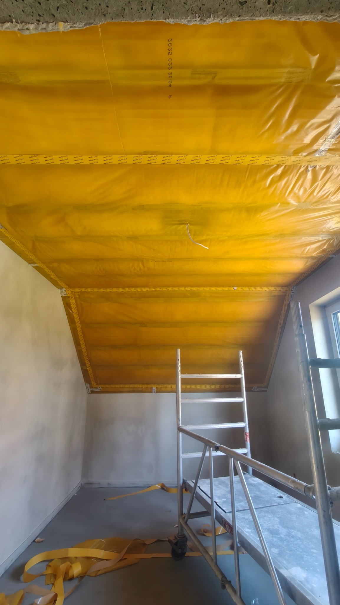 Żółta folia paroizolacyjna na suficie poddasza, zabezpieczona taśmą. Widoczne rusztowanie i fragment okna. Przygotowanie do prac wykończeniowych.