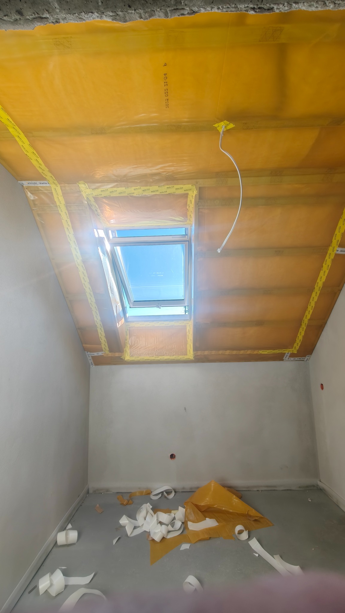 Poddasze z oknem dachowym w trakcie zabezpieczania folią i taśmą. Przygotowanie do dalszych prac wykończeniowych. Widoczne fragmenty folii na podłodze.