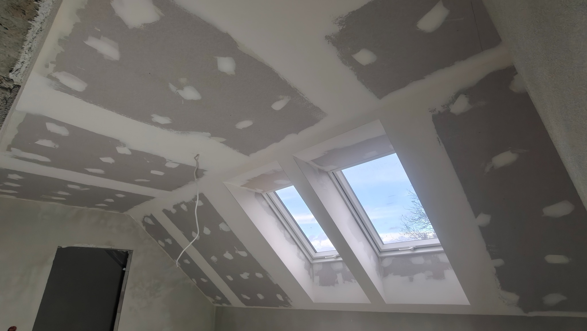 Poddasze z oknami dachowymi w trakcie prac wykończeniowych: płyty karton-gips na skosach i suficie, widoczne łączenia płyt przygotowane do szpachlowania.