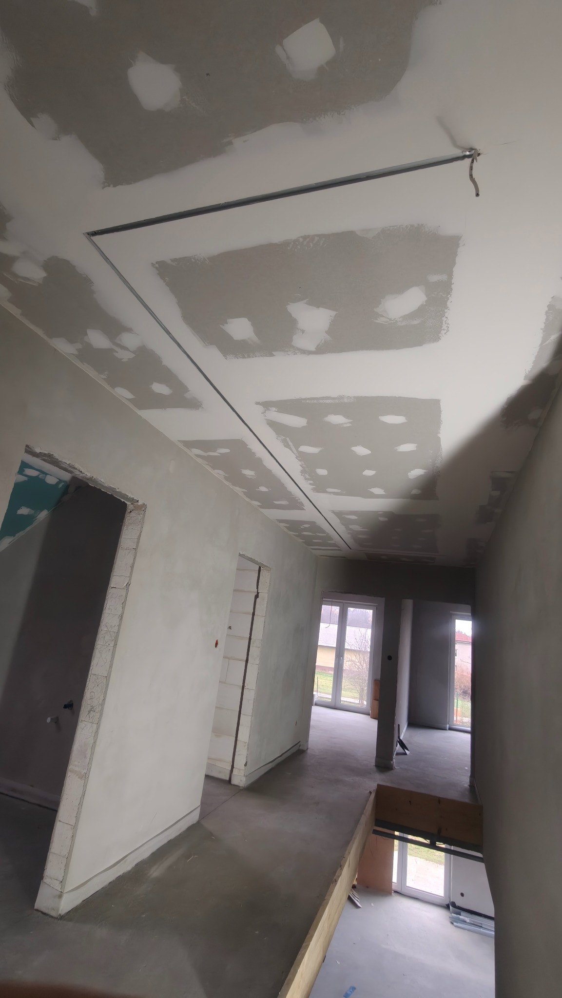 Sufit z płyt kartonowo-gipsowych w trakcie montażu, widoczne profile i szpachlowane łączenia. Surowy stan wnętrza z otworami drzwiowymi i oknami w tle.