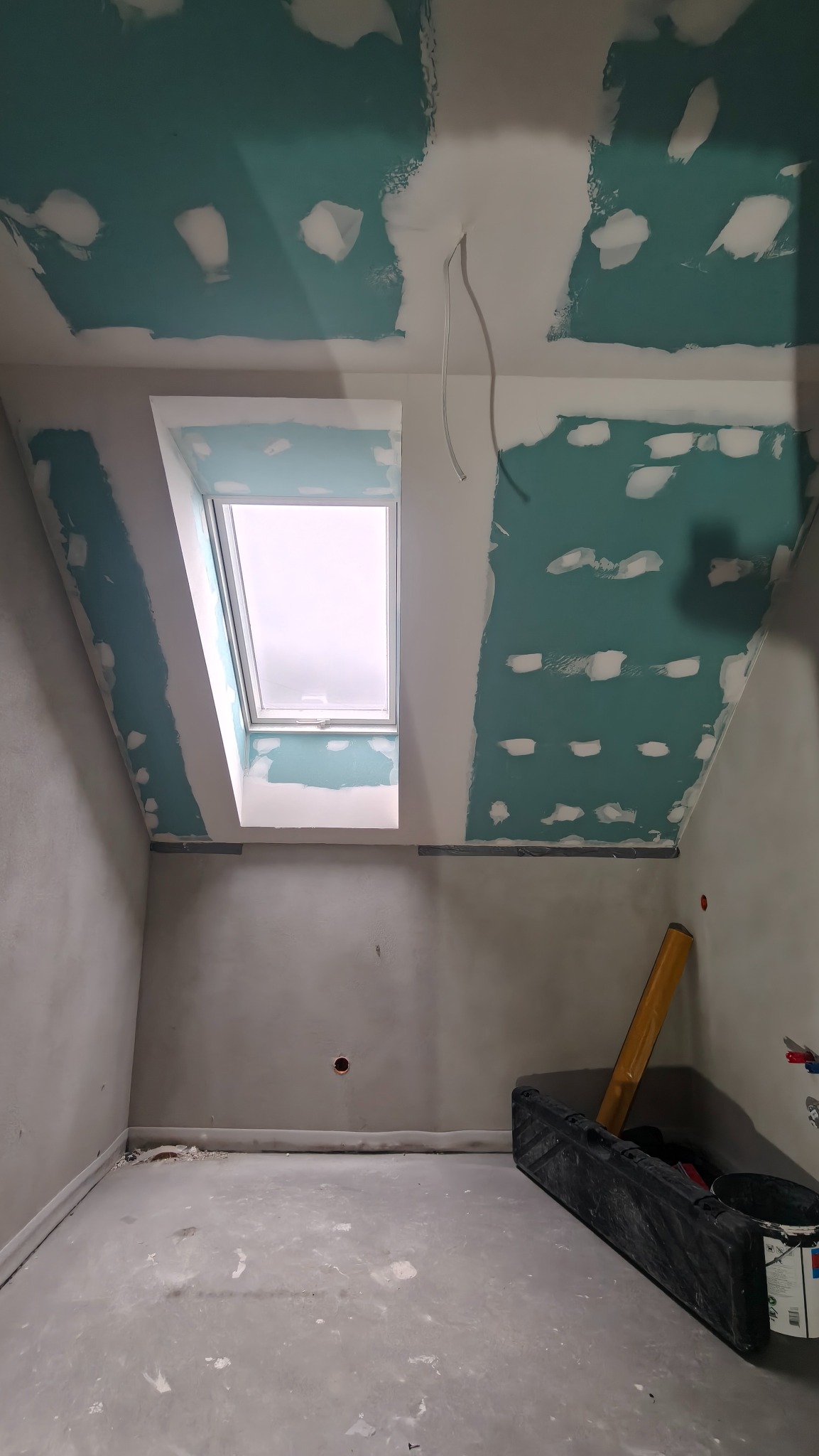 Poddasze w trakcie prac wykończeniowych: sufit i ściany z płyt gipsowo-kartonowych, częściowo pokryte szpachlą, okno dachowe, widoczne narzędzia i materiały budowlane.