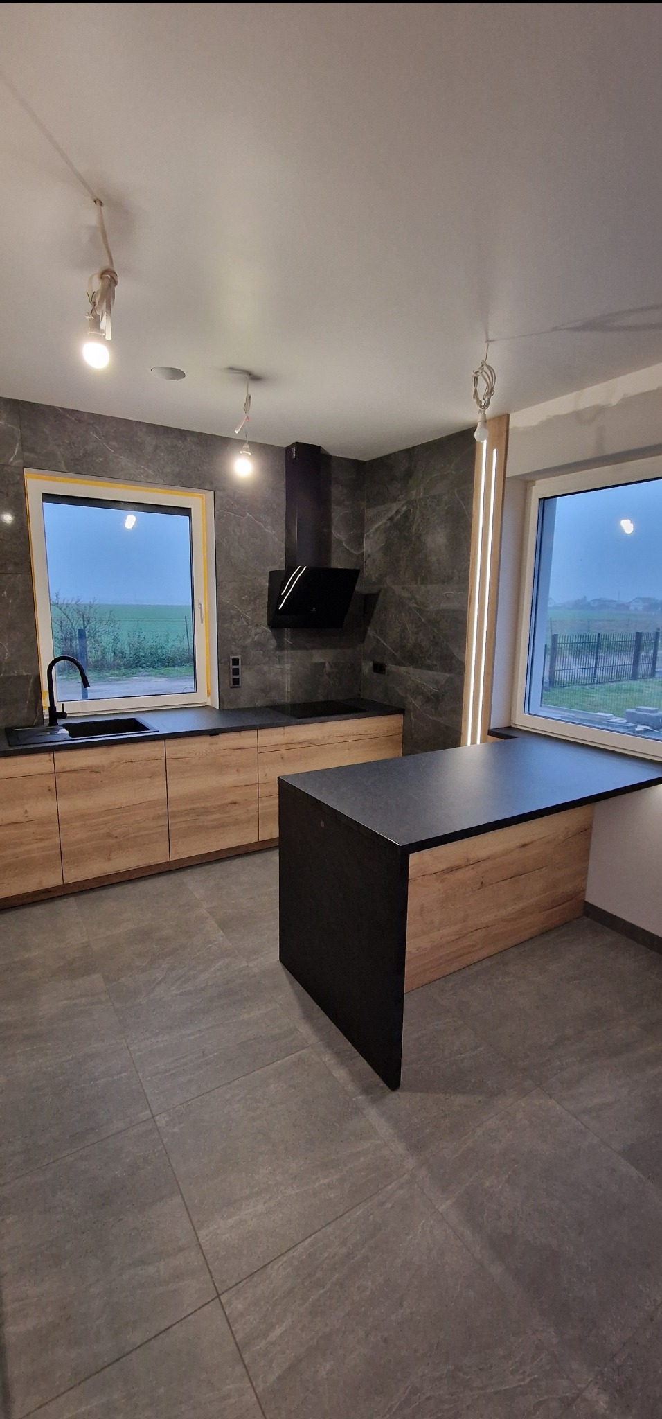 Nowoczesna kuchnia z drewnianymi szafkami i czarnym blatem, okno z widokiem na zewnątrz. Na suficie widoczne niedokończone instalacje oświetleniowe.