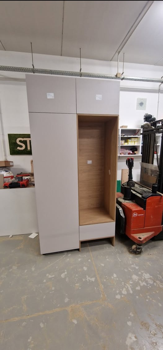 Montaż szafy wnękowej z jasnymi frontami i drewnianym wnętrzem w warsztacie, obok wózek widłowy. Widoczne elementy mocujące do sufitu.