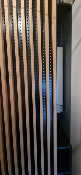 Nowoczesna zabudowa meblowa z pionowymi lamelami w odcieniach drewna i czerni, widoczny fragment szafy i biały element instalacji.