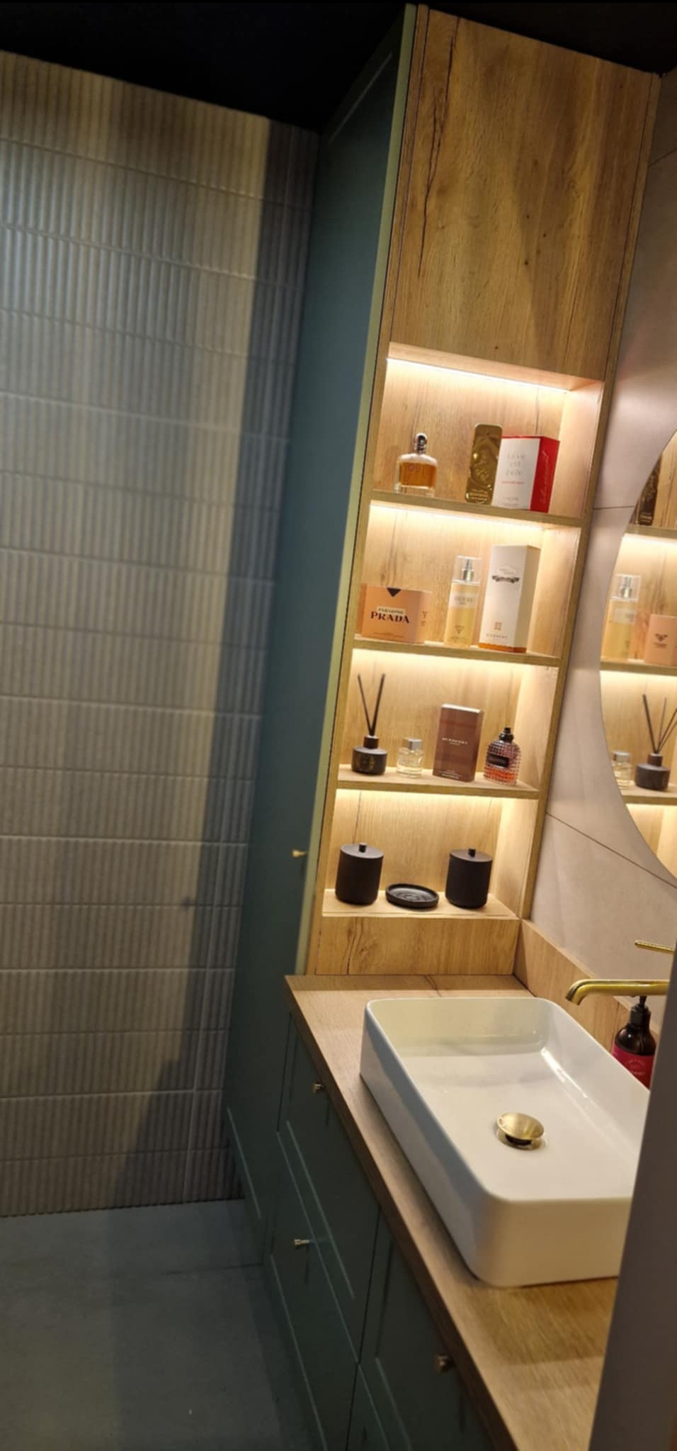 Stylowa łazienka z szafką na wymiar o drewnianym wykończeniu, podświetlanymi półkami i białą umywalką nablatową. Uwagę przykuwają flakony perfum na półkach.