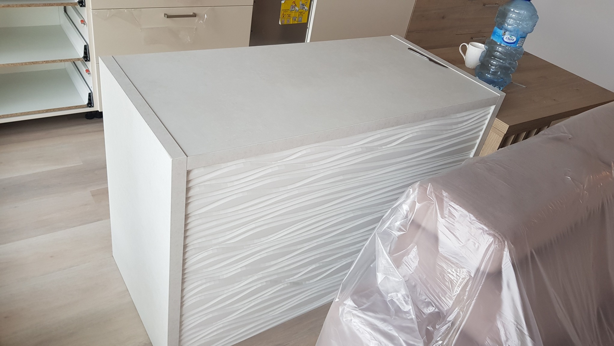 Biała szafka kuchenna z falistym wzorem na froncie, częściowo zakryta folią ochronną. Widoczne otwarte półki i blat. Elementy montażu w tle.