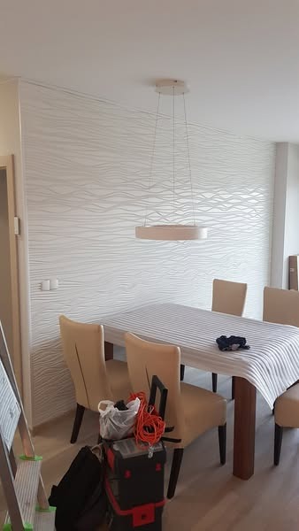 Montaż lampy wiszącej nad stołem jadalnym. Na pierwszym planie narzędzia i drabina, sugerujące trwający proces instalacji. W tle strukturalna, biała ściana.