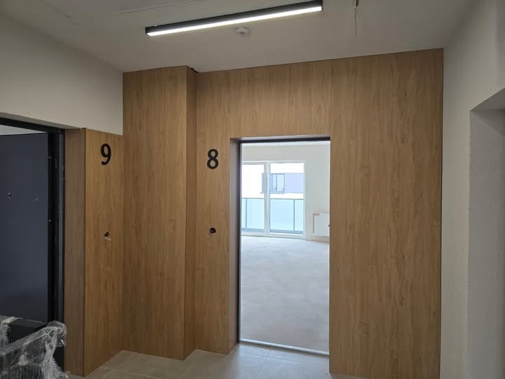 Wykończenie korytarza z numerami mieszkań 8 i 9, obłożone drewnopodobną płytą. Widoczne drzwi wejściowe i widok na balkon przez otwarte drzwi do mieszkania.