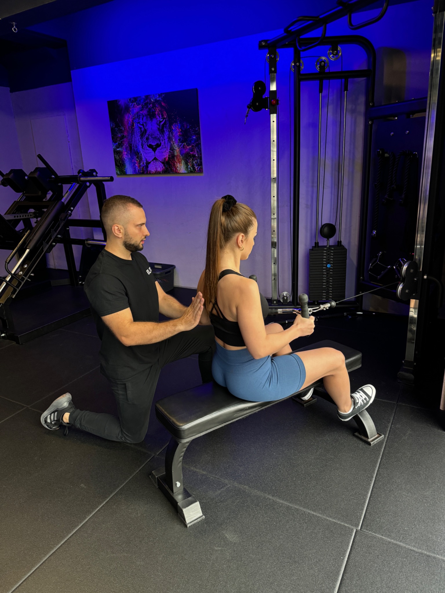Kobieta ćwiczy na maszynie pod okiem trenera w czarnym stroju. W tle ściana z neonowym obrazem lwa i profesjonalny sprzęt do ćwiczeń.